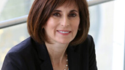 Kerry Kelly in a black sport coat leans against a wall for her headshot